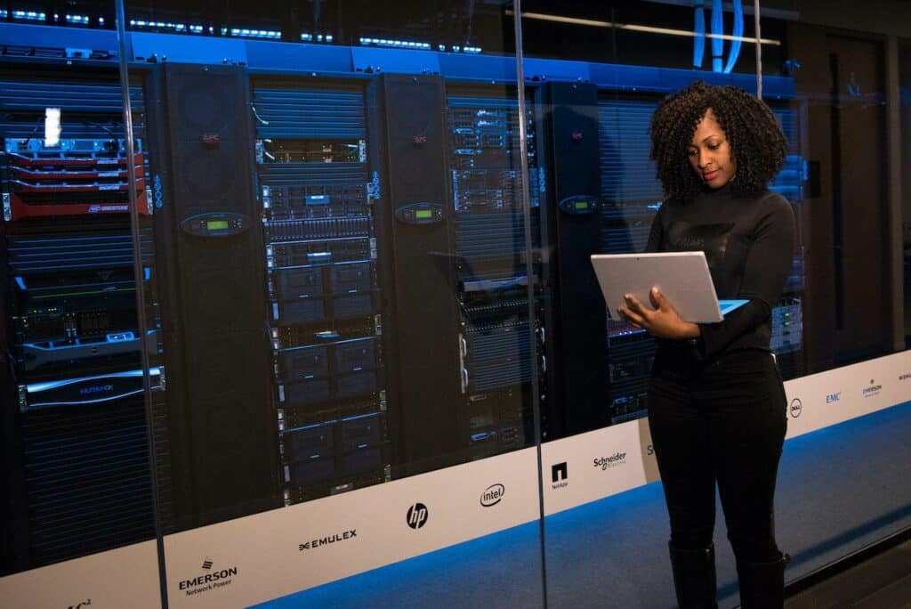 A woman stands with a laptop in her hand next to company-owned servers used for HERE Self Hosting.